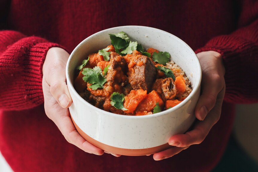 Heidi Sze's hands holding a bowl of beef, cinnamon and sweet potato stew, served over couscous for a hearty dinner for winter.