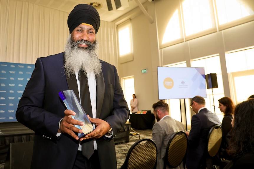 A man wearing a turban stands in a room full of people holding a trophy, smiling.