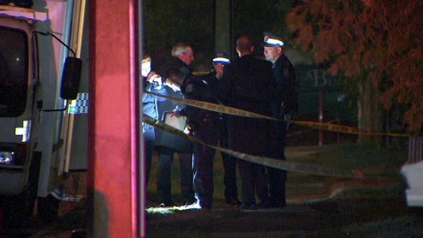 Police officers stand in a torch-lit night scene. Police tape is in the foreground.