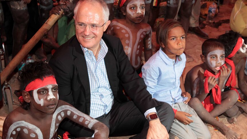 Malcolm Turnbull dressed in a suit sits on the ground with Indigenous children.