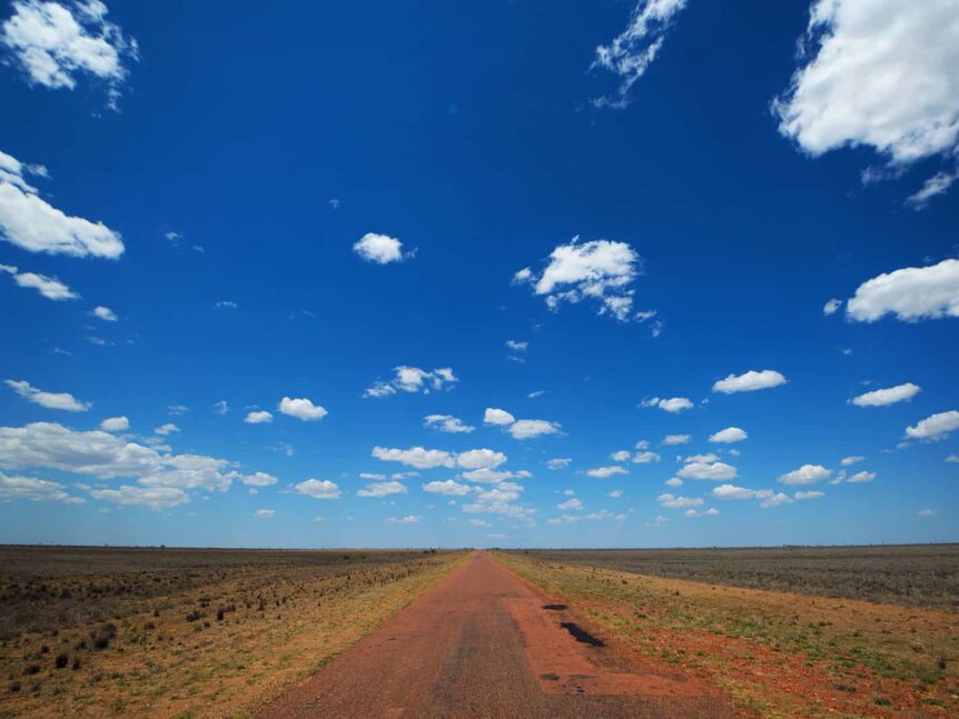 A red road juts out straight into the horizon beneath blue sky and surrounded by desert landscape.