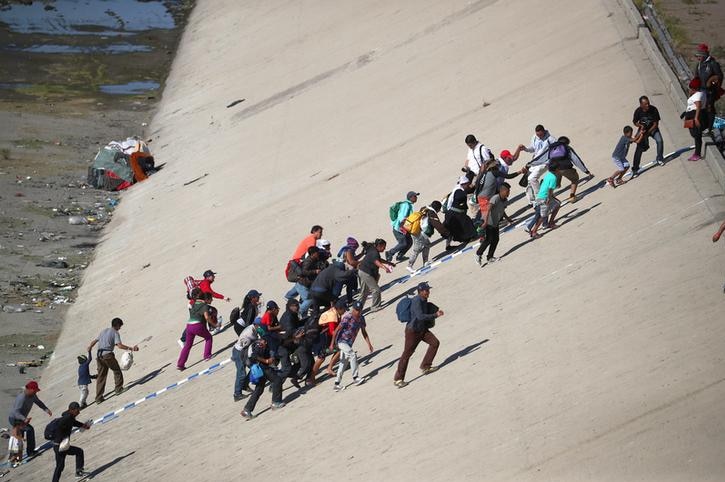 A group of migrants make their way to the border fence, lugging their personal belongings up a steep incline under harsh sun.