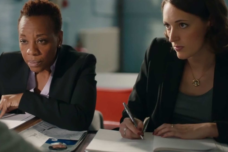 Two lawyers in suits lean against a table and stare across at an unseen person.