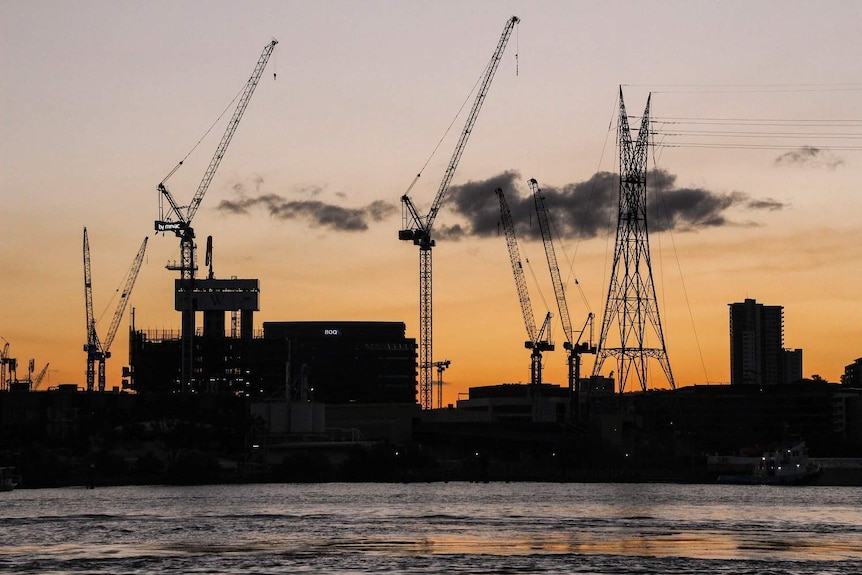 Six large cranes tower over buildings on Brisbane's skyline, taken at sunset facing Newstead from across the river