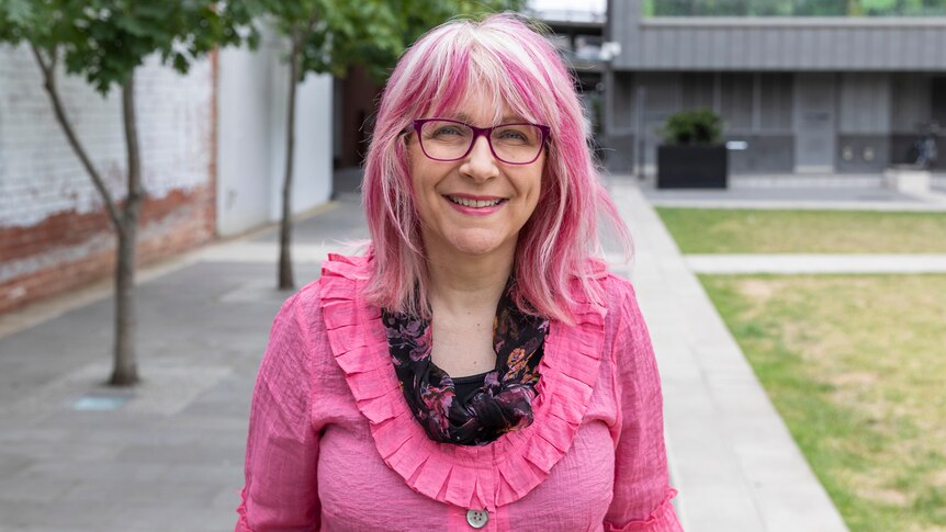 A woman with pink hair wearing a pink cardigan smiles outdoors