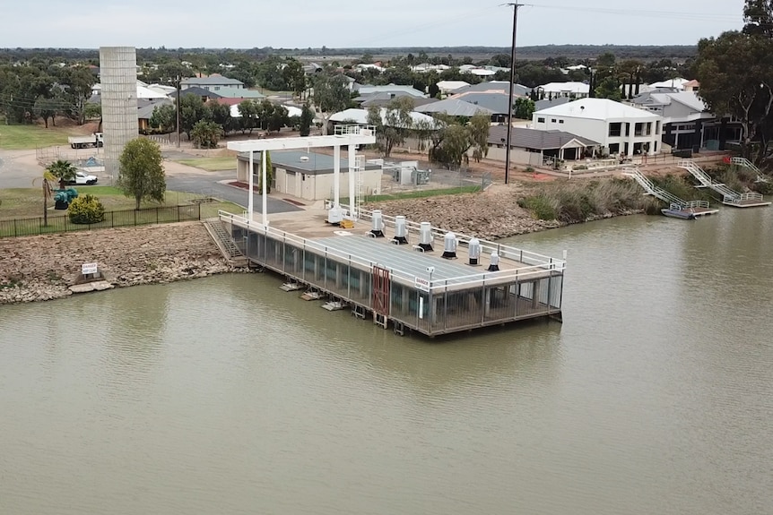 An aerial view of a pump station in a river, the sky is overcast. There are houses, a road and pump infrastructure.