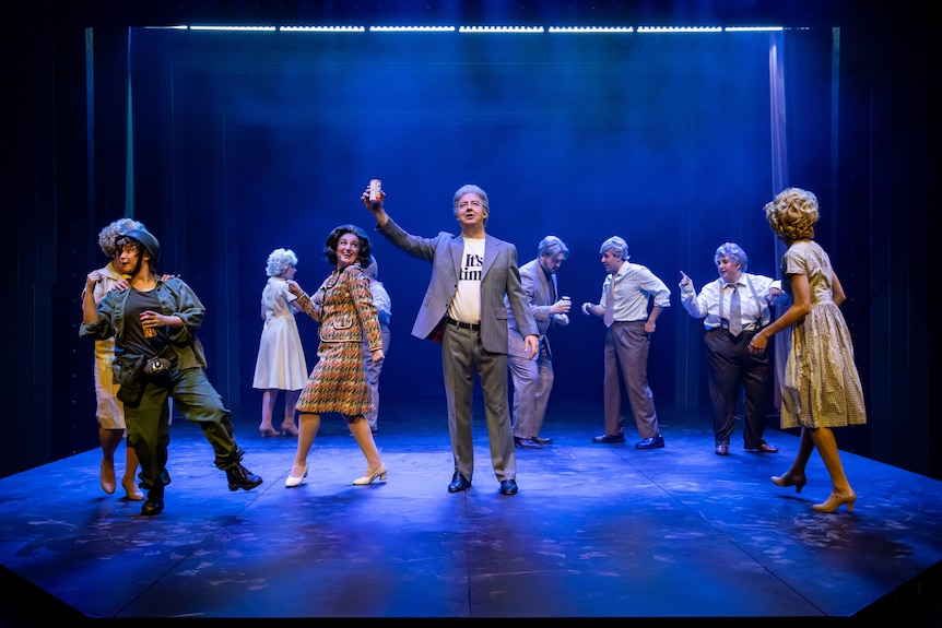 A cast of actors dressed in 70s clothing sing and dance on a blue-lit stage. In the centre is Gough Whitlam holding up a beer.