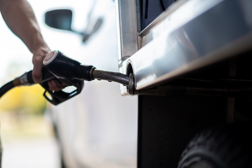 A close-up of a man's arm refuelling a white ute with a petrol bowser.
