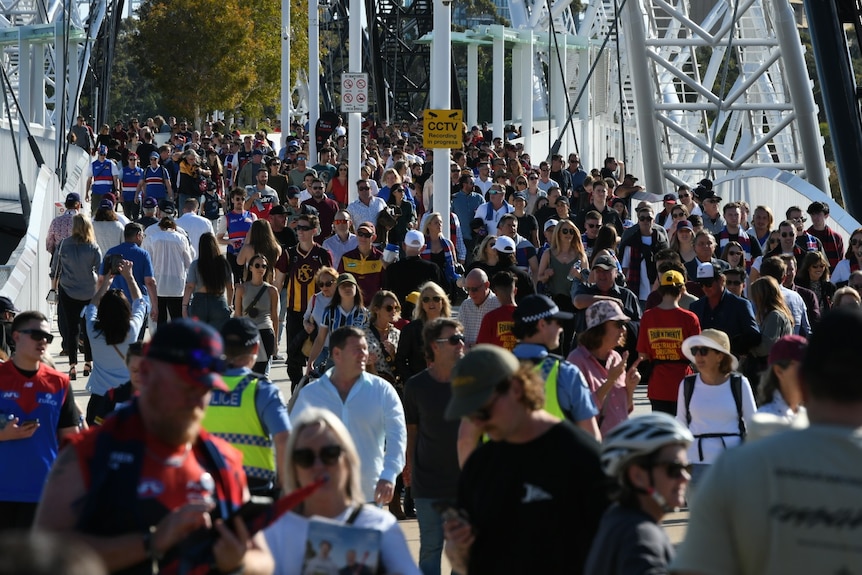 Hundreds of people in AFL merchandise stream across Matagarup Bridge on a sunny day.