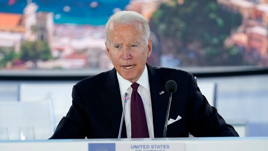 Joe Biden sits at a conference desk, with a sign saying "United States of America"