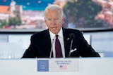 Joe Biden sits at a conference desk, with a sign saying "United States of America"