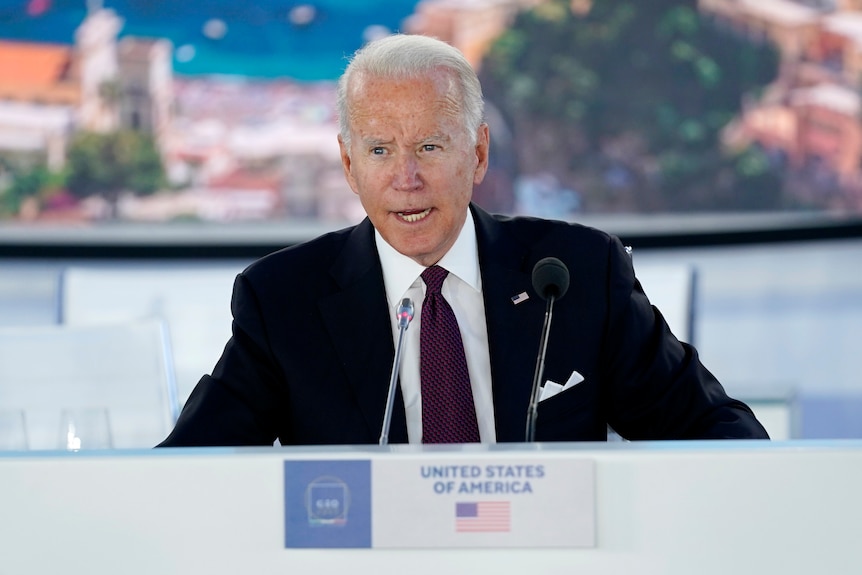 Joe Biden sits at a conference desk, with a sign saying "United States of America"