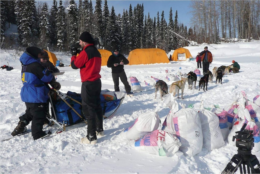 Sled dogs, tents and people filming in a snowy landscape