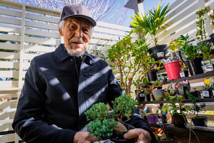 Ziggy stands, smiling, holding a bonsai plant, in front of a row of plants in his outdoor garden space.