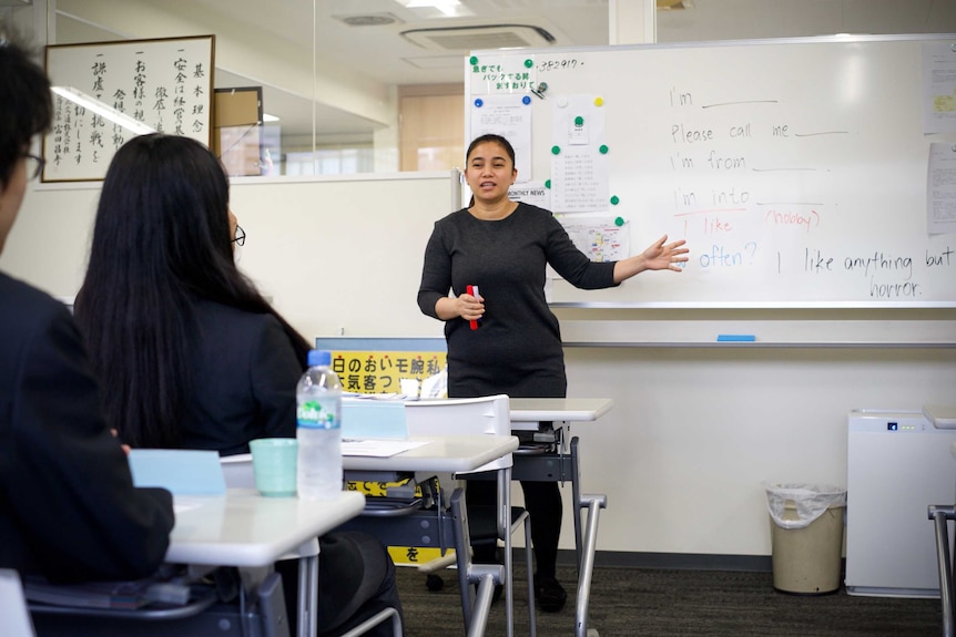 A woman stands in front of a classroom with English phrases on a whiteboard