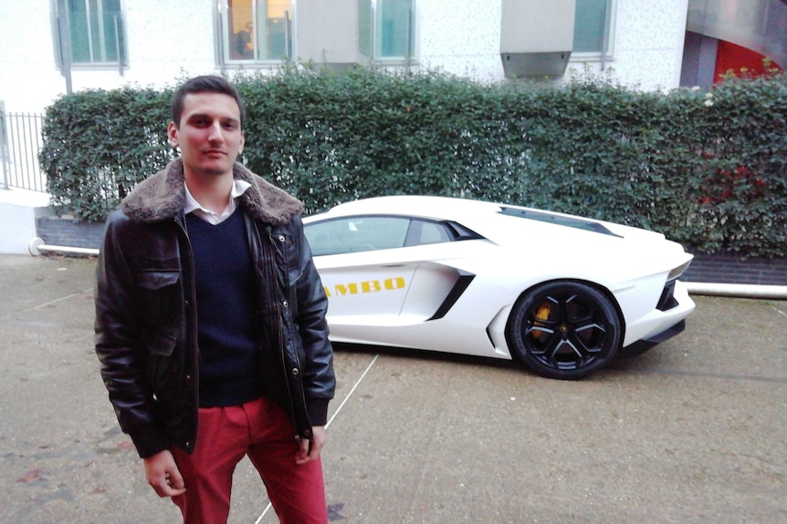 A man in a leather jacket stands in front of a sports car.