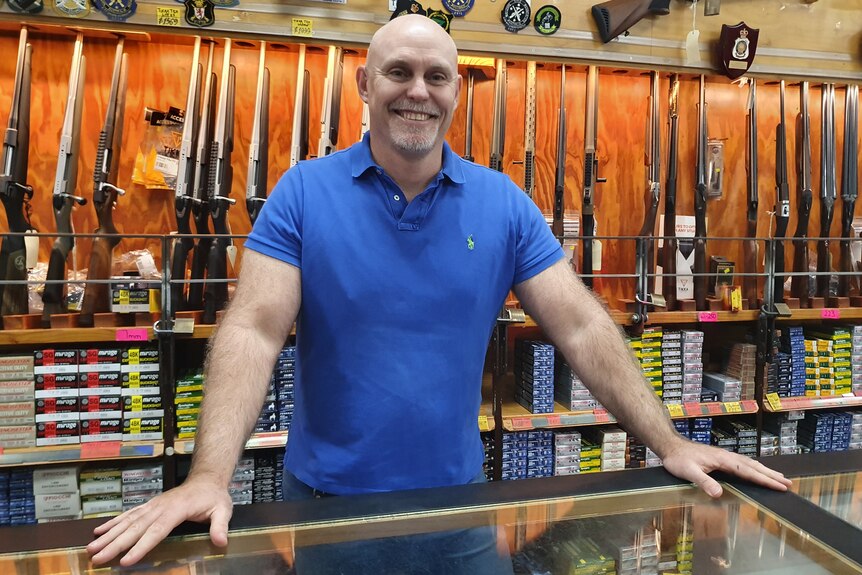 Owner of gun shop standing at counter with line of guns on wall behind him