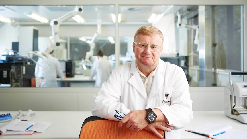 Dr Nicholson stands in front of two people working in a lab behind him. He wears a white lab coat with "Murdoch University".