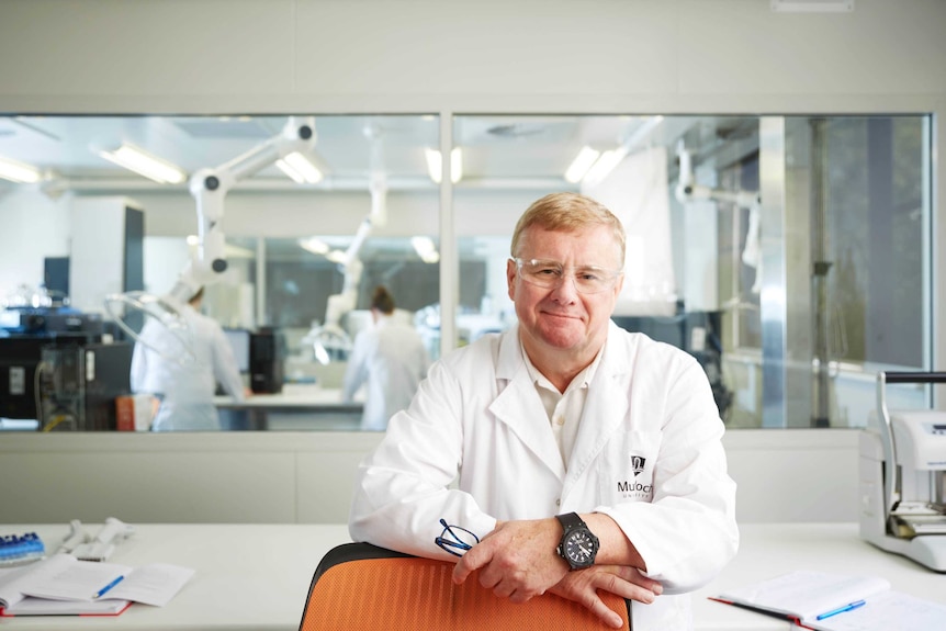 Dr Nicholson stands in front of two people working in a lab behind him. He wears a white lab coat with "Murdoch University".