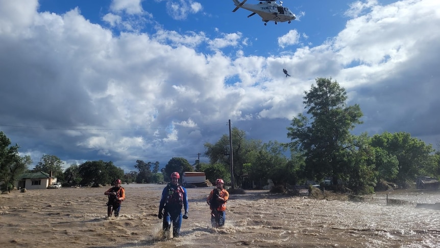 Three people walk through floodwater while a helicopter winches someone into the air behind them.