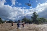 Three people walk through floodwater while a helicopter winches someone into the air behind them.