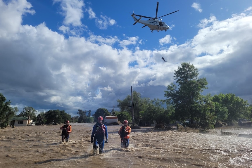 Three people in rescue team clothes walk through floodwater. There is a helicopter in the sky.