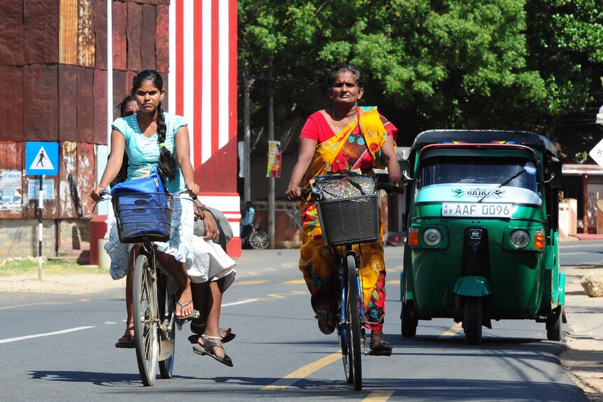 Two women in orange and blue saris ride bicycles, a green tuktuk follows.