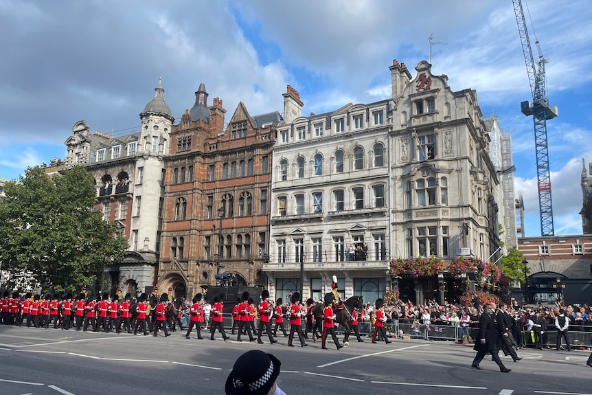 A band dressed in royal uniforms with red jackets marching down a London road with historic buildings in the background