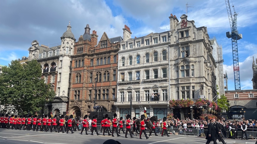 A band dressed in royal uniforms with red jackets marching down a London road with historic buildings in the background