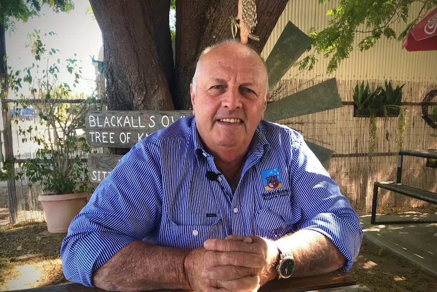 A friendly looking, older bald fellow in a blue shirt sits at what looks like a cafe table.