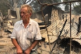 A man in a white shirt covered in ash stands in front of twisted, burnt metal wreckage.