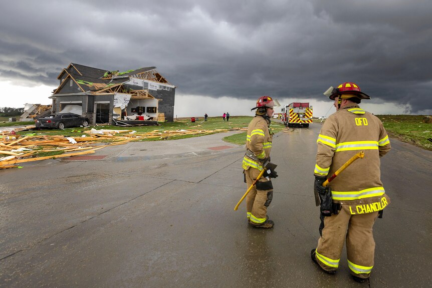 Firefighters stand in front of a damaged home with dark storm clouds in the background. 