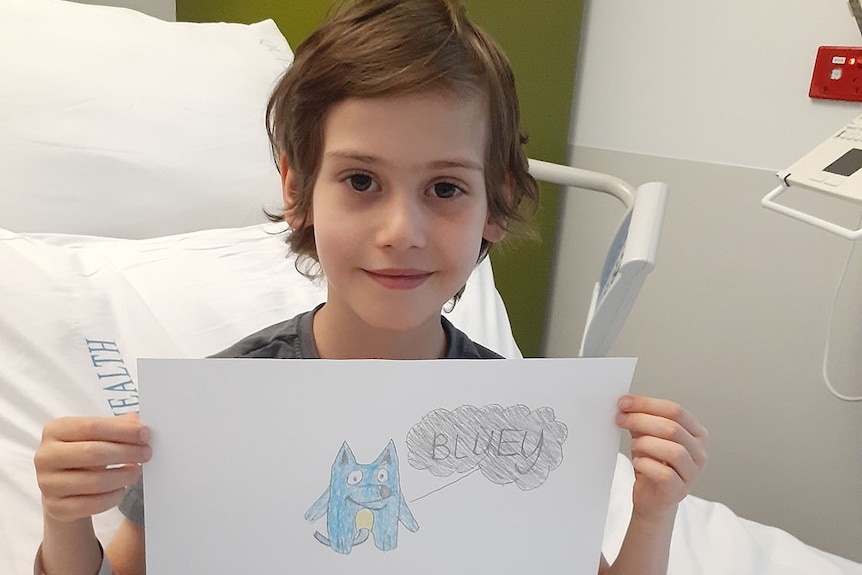 Zen holds up a drawing of Bluey from a hospital bed.