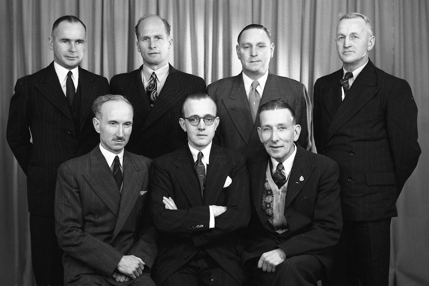 Historic image shows seven men in suits posed for informal indoor staff photograph.