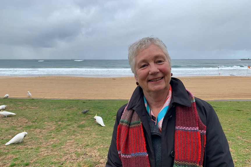 A woman, wearing a jacket and scarf, stands at a beach near Wollongong