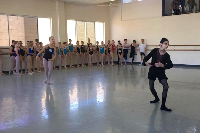 A ballet teacher instructs a class of dancers in a hall