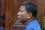 A man stands in a blue shirt in a Vietnamese wooden court room speaking into microphone, guards in background.