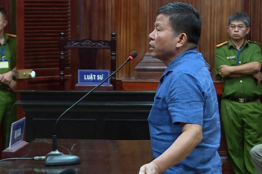 A man stands in a blue shirt in a Vietnamese wooden court room speaking into microphone, guards in background.