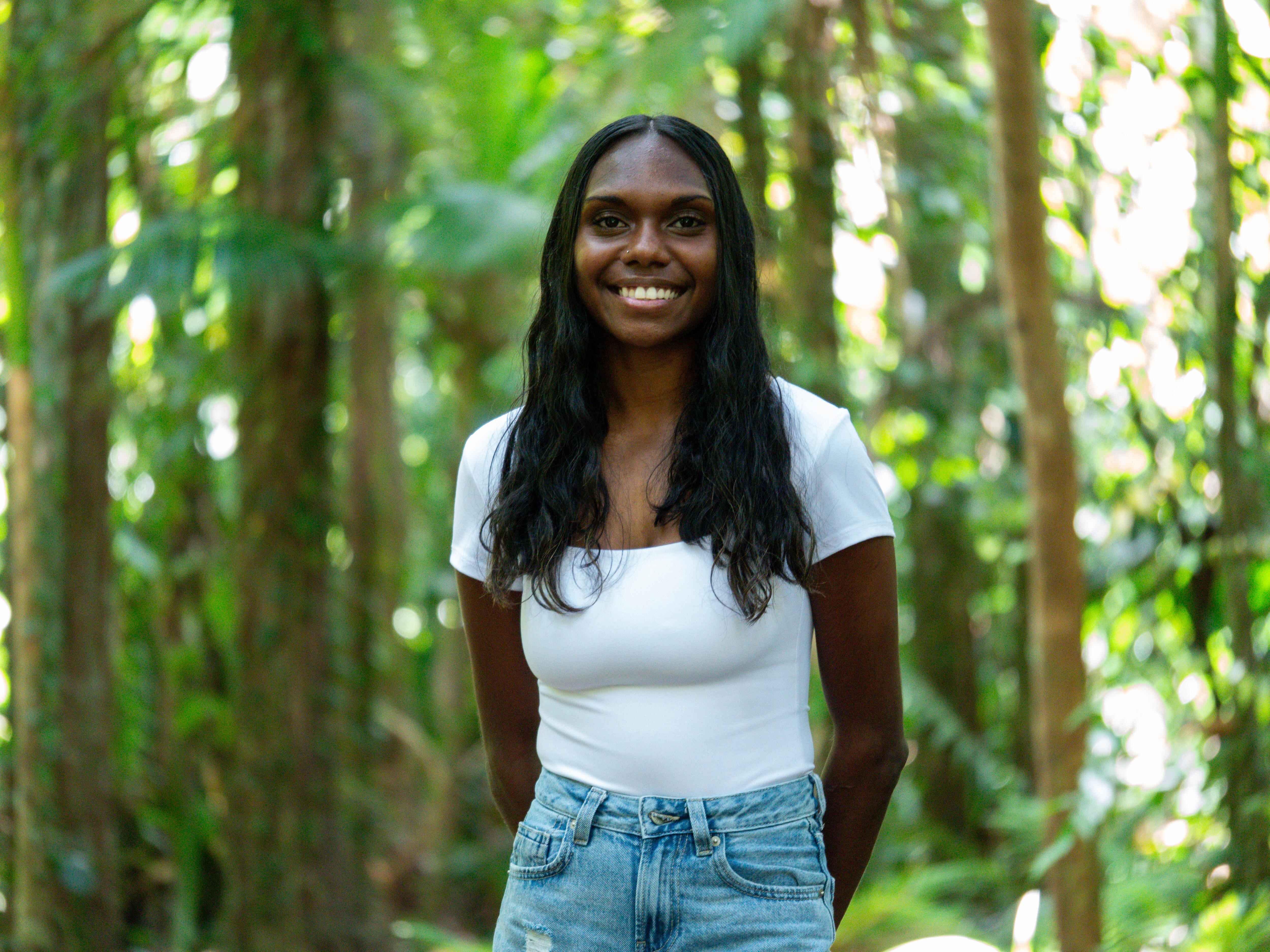 A young Black woman with black hair wearing a white t-shirt and blue jeans smiles at the camera, behind her are green trees