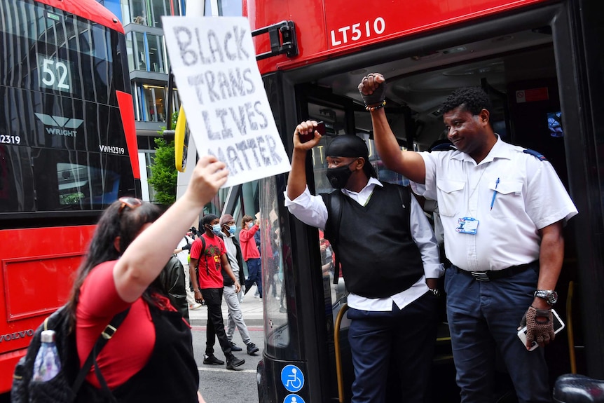 You view an image of red London buses stopped as bus drivers in an open door raise their brown fists in solidarity with marchers