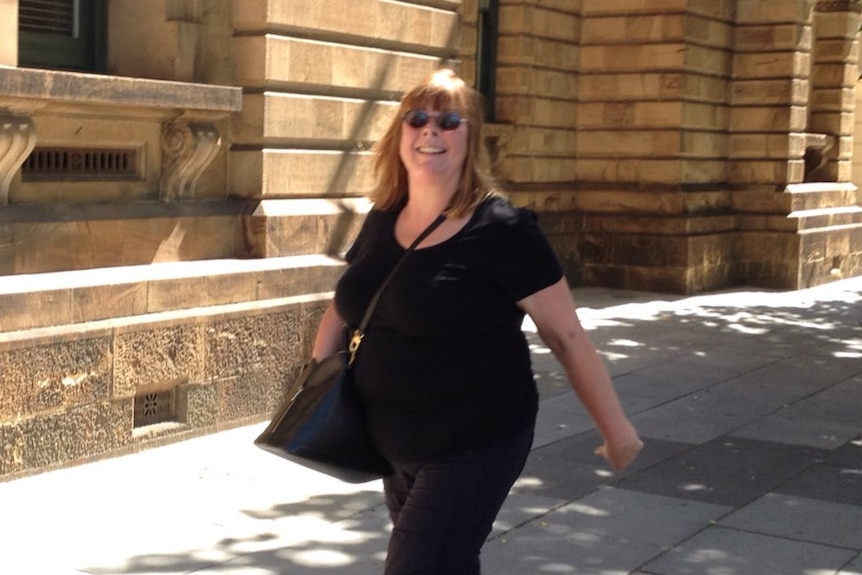 A woman with brown hair wearing all black and sunglasses walks past a stone building