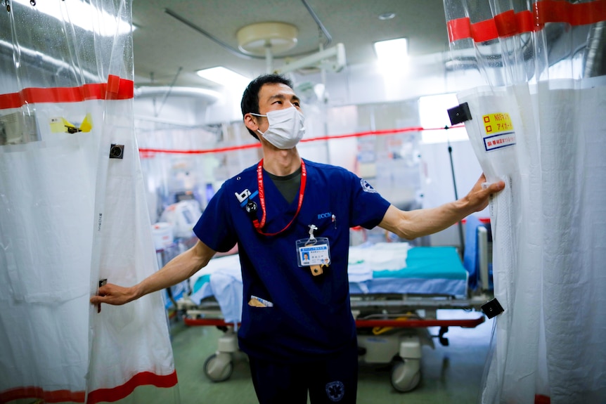 A male doctor in blue scrubs and a mask reaches to close curtains in a hospital ward