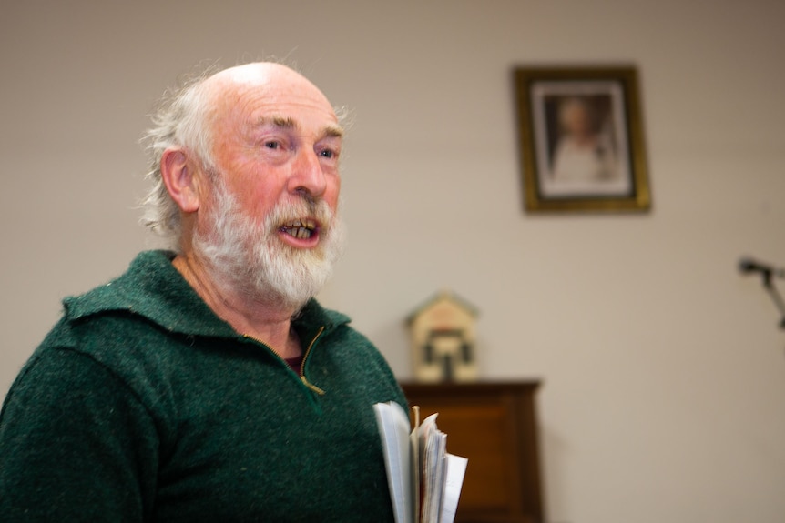 An ageing man speaking to an unseen audience in a small town hall.