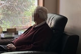 An older man wearing a maroon jumper looks out the window, away from the camera.