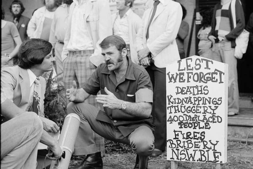 A man with a moustache kneeling next to a mock tombstone and talking to a reporter.