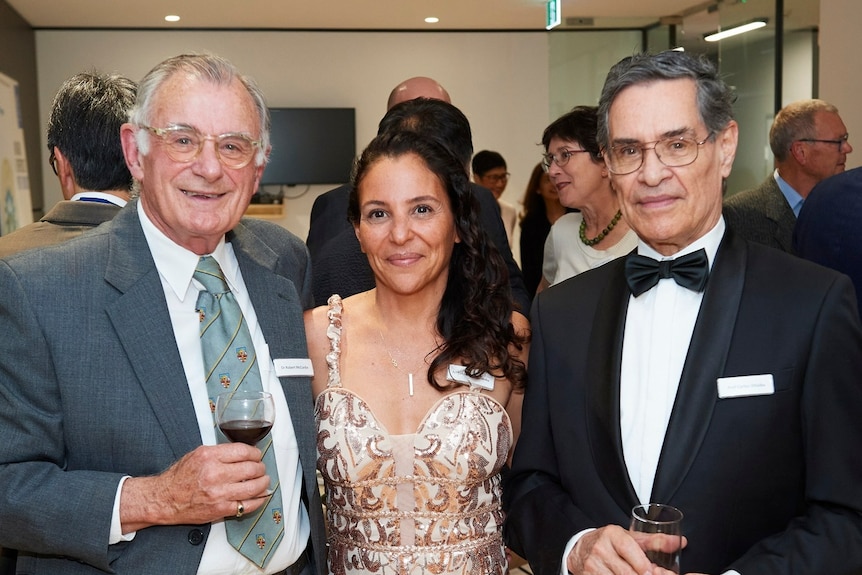 Woman in glittery dress smiling between two men in suits, one holding wine glass