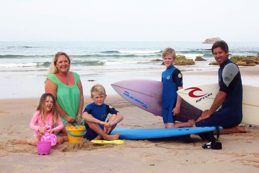 A man and woman sitting on a beach, with two young boys and a girl, and surfboards between them.