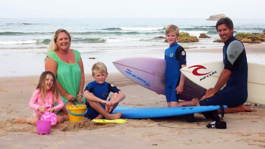 A man and woman sitting on a beach, with two young boys and a girl, and surfboards between them.