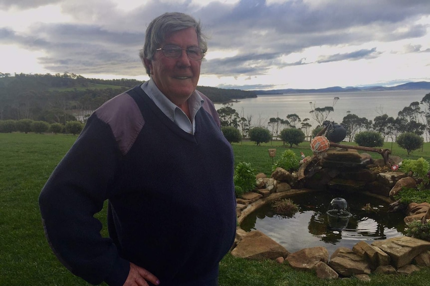 Man with white hair stands before view of calm ocean on overcast day