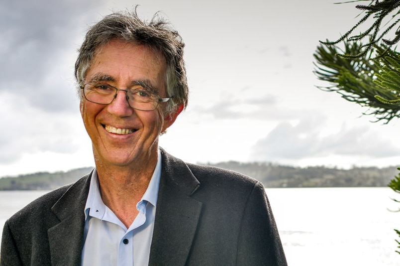 A smiling, grey-haired bespectacled man in an open-necked blue shirt and dark blazer stands in front of a body of water